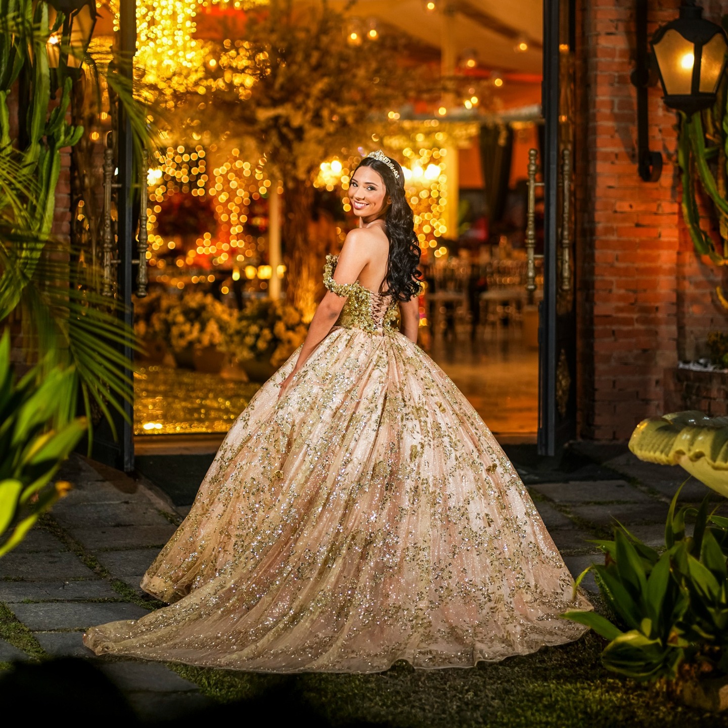 Debutante em vestido dourado olhando para trás em entrada de tijolinhos decorada com luzes