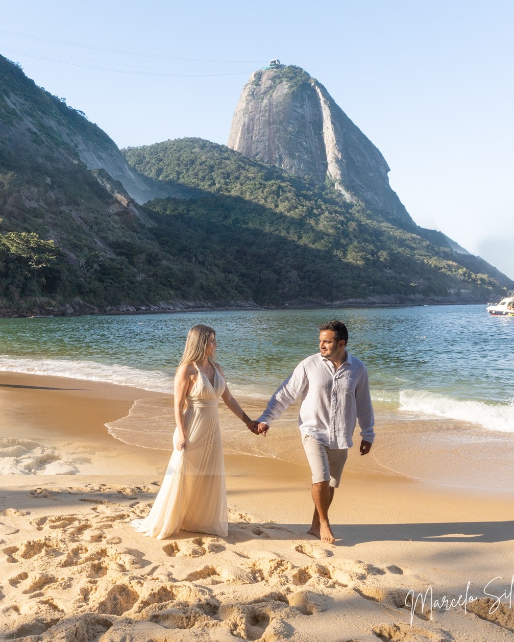 Noivos de mãos dadas caminhando na praia com o Pão de Açúcar ao fundo