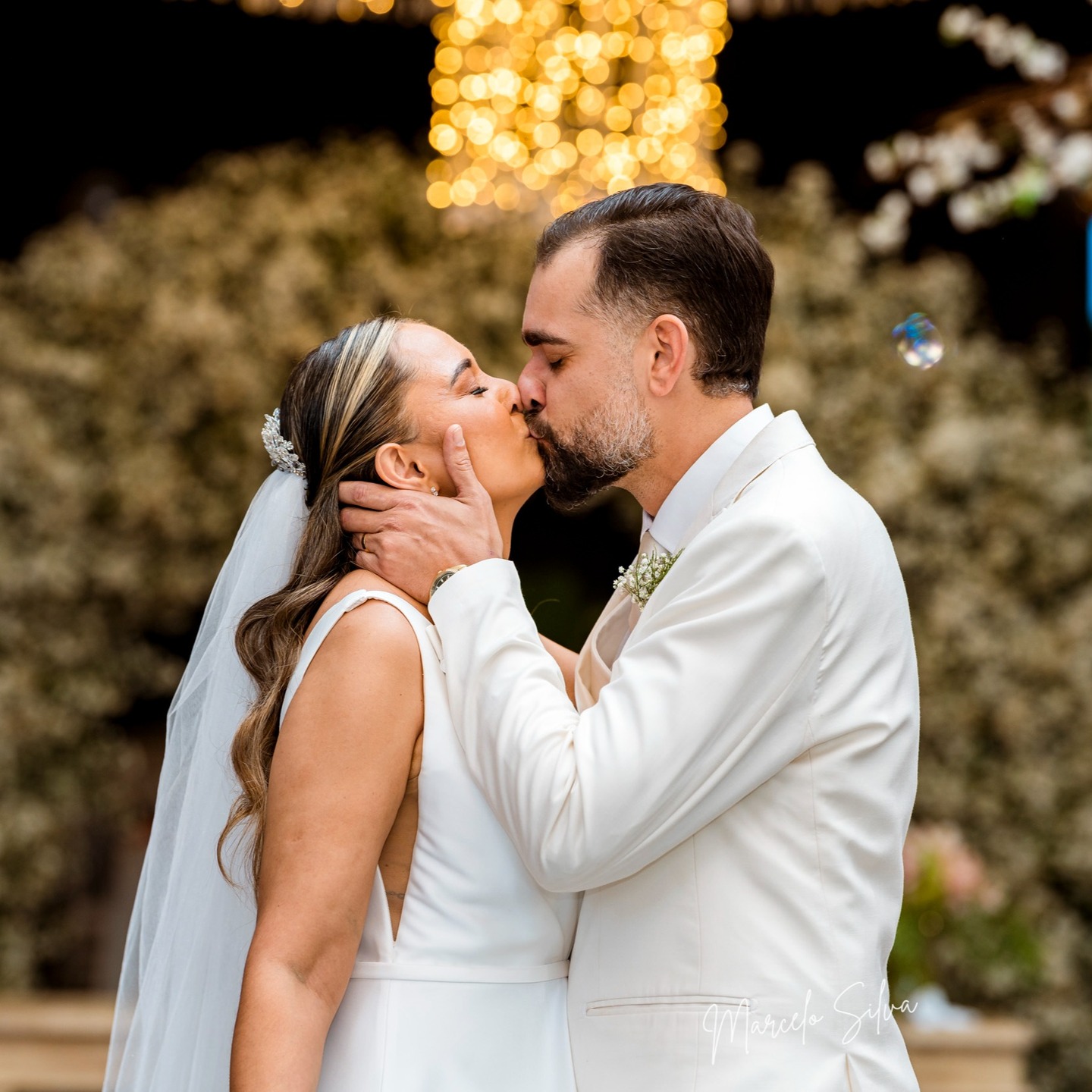 Noivos se beijando em jardim florido iluminado por luzes suspensas